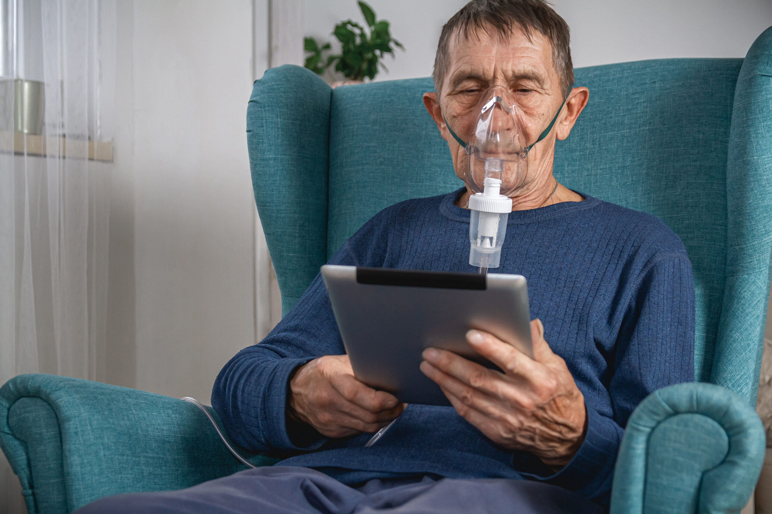 An elderly senior sits in a armchair with an oxygen mask while getting pulmonary rehab at home